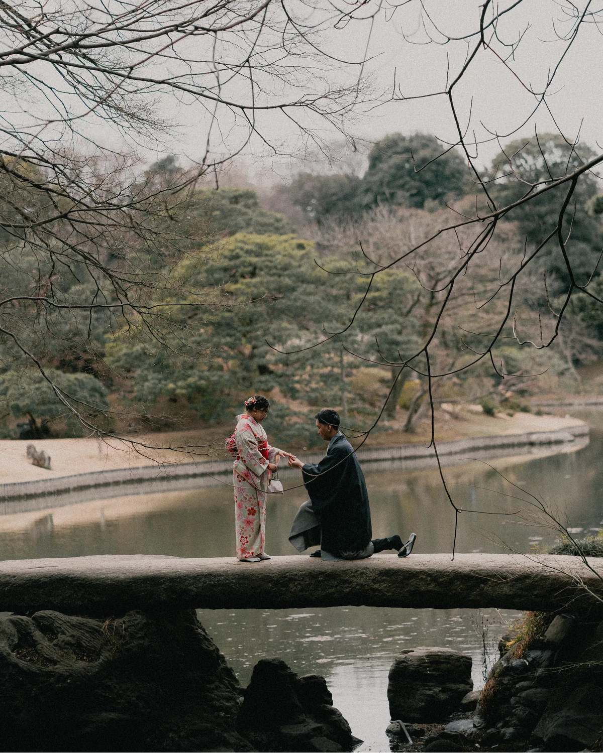 A couple in traditional Japanese attire stands on a stone bridge over a pond, framed by bare tree branches overhead. The woman wears a light pink floral yukata, and the man kneels in a dark kimono.
