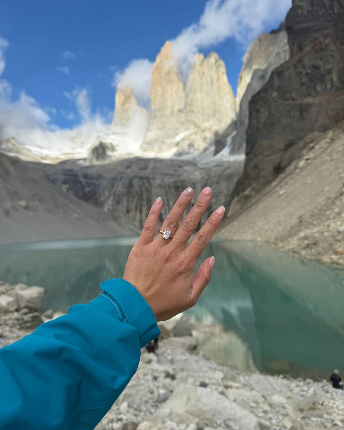 A person's hand wearing a old mine cut 18k yellow gold diamond engagement ring, set against a stunning background of a glacial lake and towering mountain peaks under a blue sky.