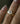 "Close-up of a pear-cut diamond engagement ring in 18k yellow gold, featuring a large brilliant diamond set in a sculptural flowing setting, shown on a person's finger with sparkly nail polish against a dark background.