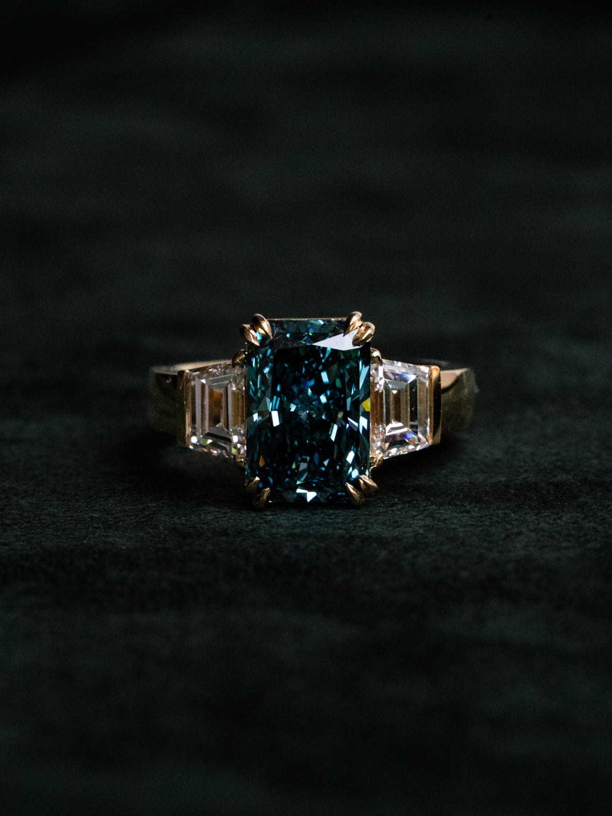 Fancy vivid blue radiant cut diamond ring in 18k yellow gold with trapezoid side stones, shot on dark velvet